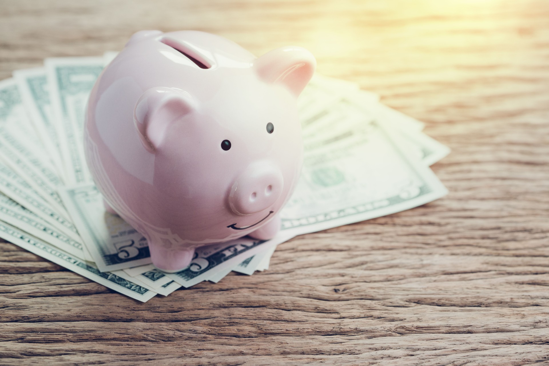 Pink piggy bank on pile of US Dollar banknotes on wooden table with artificial golden flare light, finance, banking, saving money investment concept